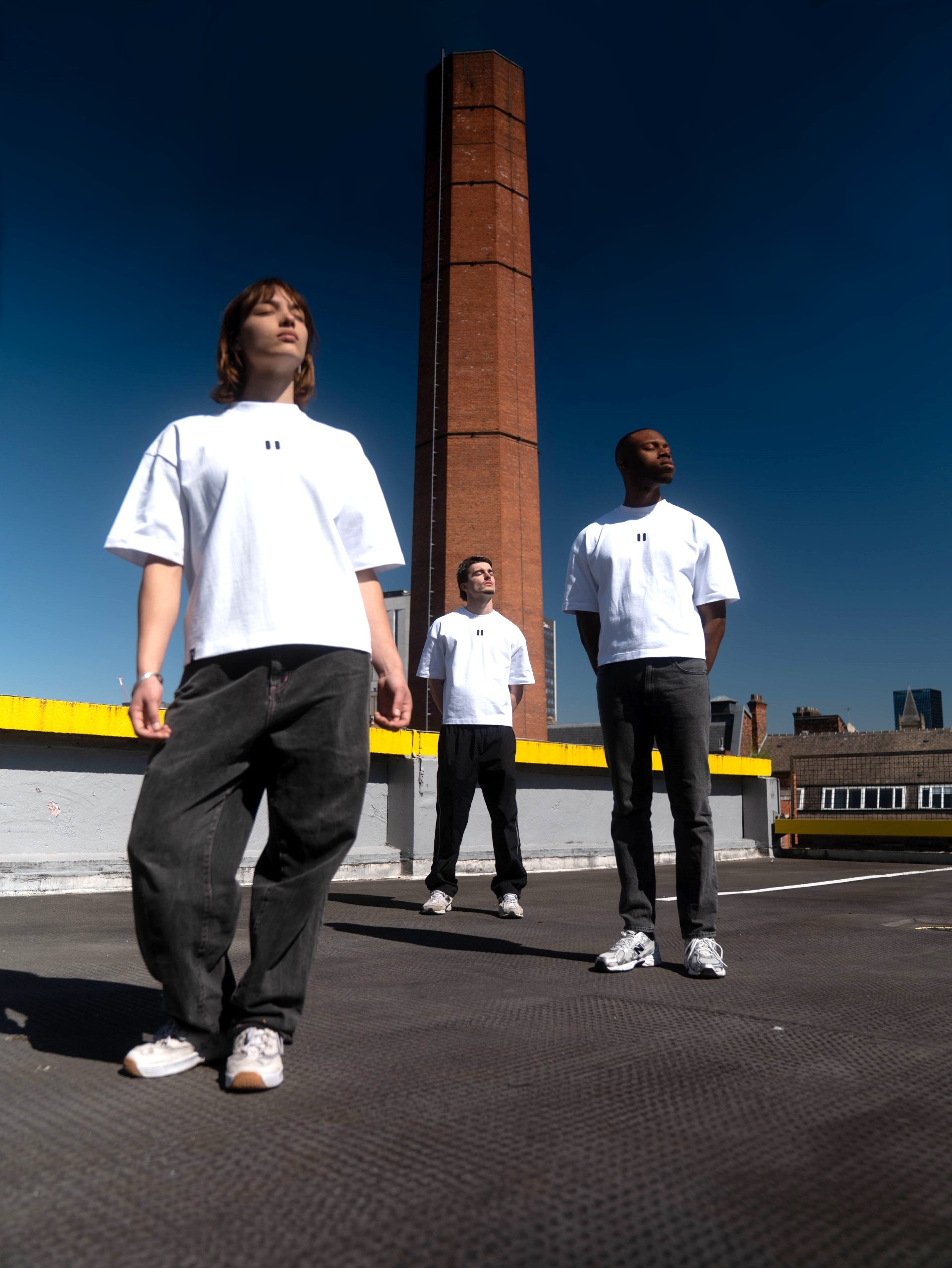 Three people wearing white t-shirts standing on a rooftop with a tall brick tower in the background.